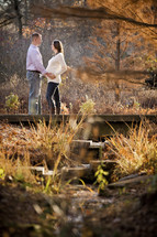 man with his hands on the belly of his pregnant wife standing on a small bridge on a nature trail