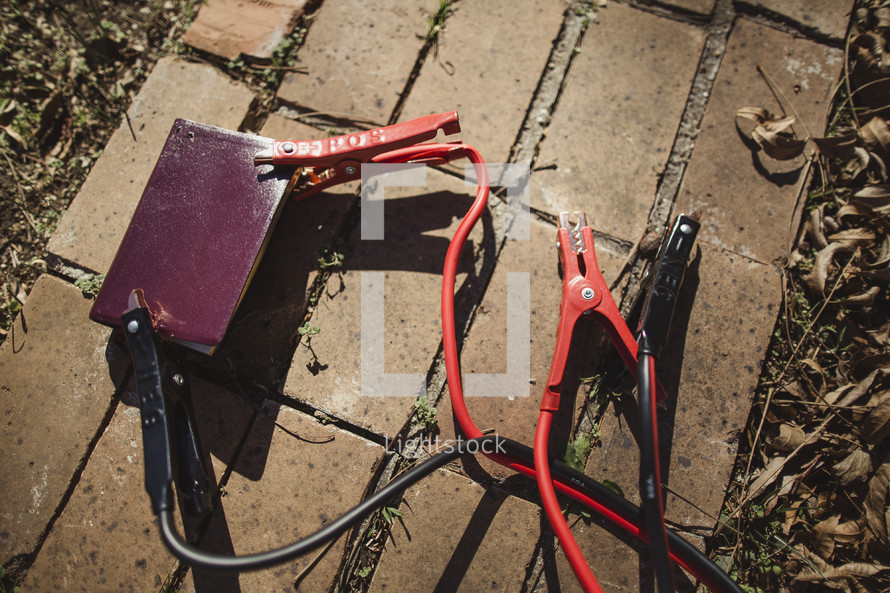Bible laying on brick sidewalk with jumper cables attached.