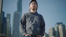 A contemplative young man walks along a dock towards the camera with a city skyline in the background