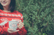 a woman holding a mug of hot chocolate standing in front of a Christmas tree 