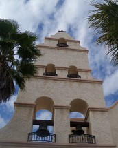 This church bell tower reaches high in the sky towering above a city square reaching high into the Heavens showing the light of the world to all who pass by. This church is a new church building designed after the old spanish style missions out west.