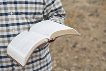 a man in a plaid shirt reading a Bible outdoors 