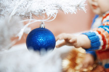 toddler reaching for blue ornament on a white Christmas tree 