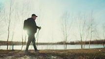 Active handsome elderly man with nordic walking sticks walks forward through along the lake at sunset. Senior man is engaged in scandinavian Walking with a backpack. Healthy lifestyle concept.