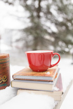 red mug on a stack of books on a table outdoors in the snow 