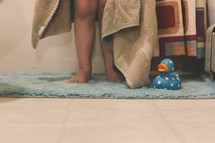 Child's legs standing on a rug with a towel and a rubber duck.
