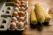 Eggs in a carton and corn on the cob on a wet cutting board.