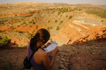 a woman sitting outdoors at the top of a canyon at sunset looking at a map