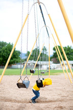 Little boy in yellow rain coat playing on the swings 