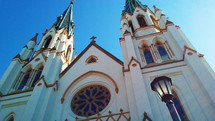 An old historic catholic church in Savannah, Georgia with large stained glass windows and gas lights stretch out into the blue sky with high arches and old gothic style architecture in the heart of the city where old and young alike can come and worship God together. 