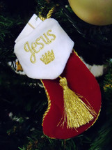 A red and white Christmas stocking with Jesus embroidered on the stocking in gold embroidery lettering waits for Jesus on a Christmas Tree surrounded by Christmas tree ornaments. 