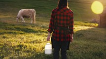 Woman stands in meadow of farm with pasture for cow with a milk can in hand for milking cattle. Agriculture animal husbandry for production of natural, healthy products. Summer sunlight on nature.