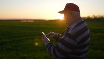 Old senior man farmer with smartphone working a in field. Senior farmer is engaged in smart farming agriculture. The tractor is running in the background.