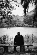 a senior man sitting alone on a bench looking out at a pond 