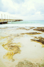 Water and mud flowing onto wet beach with pier and clouds in the background.