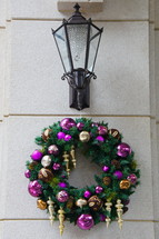 Purple and gold ornaments on pine wreath hanging under coach lamp on stone wall.