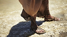 Jesus standing near the water with his feet in sandals.