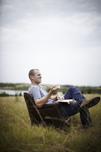 man reading a book in a leather chair sitting in a field