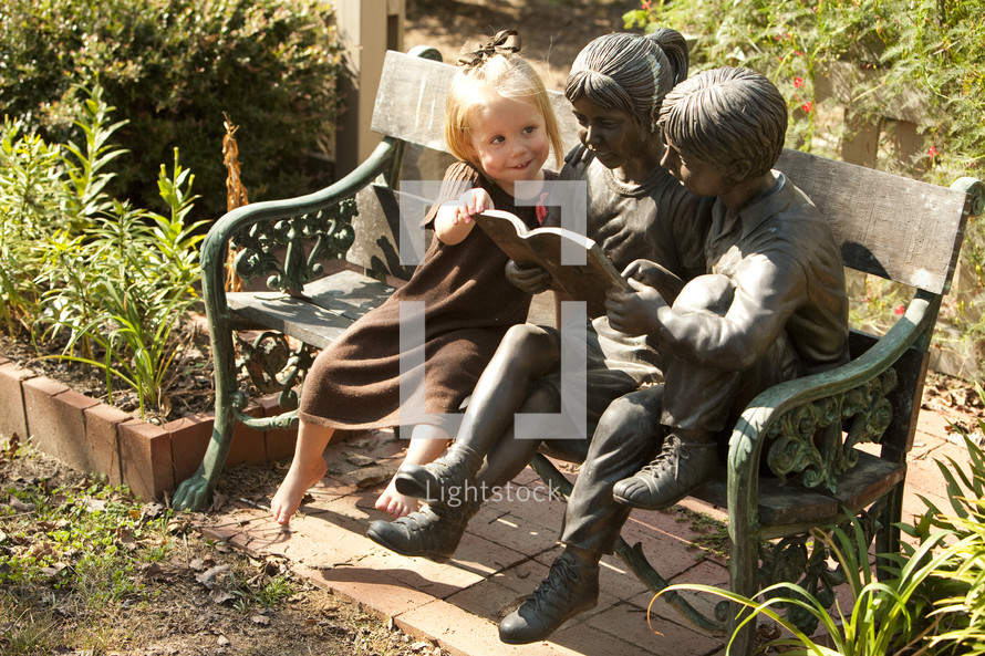A young girl reading a book with statues on a bench