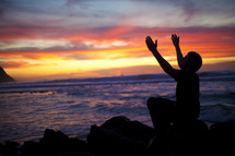man sitting on a rock by the ocean with his arms raised in worship to God during a sunset