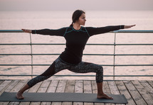 Young brunette hispanic woman doing yoga exercises on wooden sea embankment in the morning. Girl in black sports costume. Stretching, practice, healthy lifestyle concept .