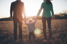 Backside view of man and woman holding the hands of a toddler child while standing in grassy field at sunrise.