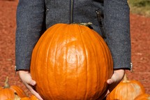 Child in gray wool jacket holding large pumpkin.