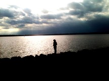 A silhouetted woman stands alone on the shore overlooking a peaceful lake with the sunlight reflecting in the water. 