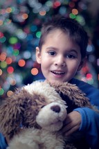 Little boy snuggling a stuffed animal  in front of a Christmas tree