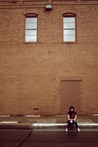 Girl sitting on a sidewalk in front of a brick building