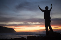 man standing in front of the ocean at sunset with hands raised in praise and worship to God