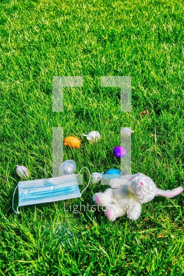 face mask, stuffed animal, and Easter eggs scattered in the grass 