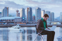 Man sitting on a chair with his laptop near and ocean bay by a city skyline.