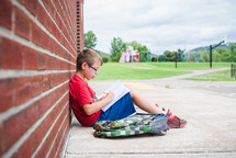 a young boy leaning against a school building doing his homework 
