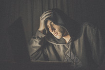 A tired man in a hooded sweatshirt sits before a laptop computer.