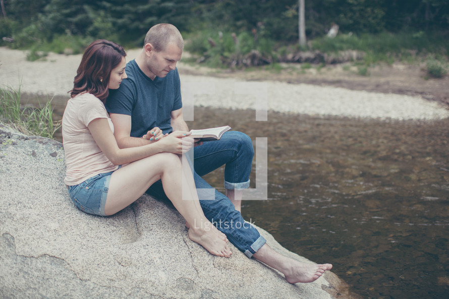 man and woman sitting on a rock reading a Bible by a river 