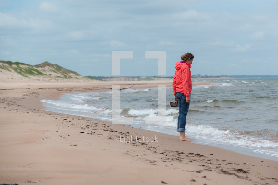 woman holding her shoes standing on a beach 
