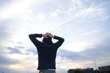 A person stands looking at the sky with hands behind his head.