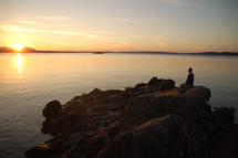 Silhouette of a man sitting on a rock formation in the ocean water at sunset.