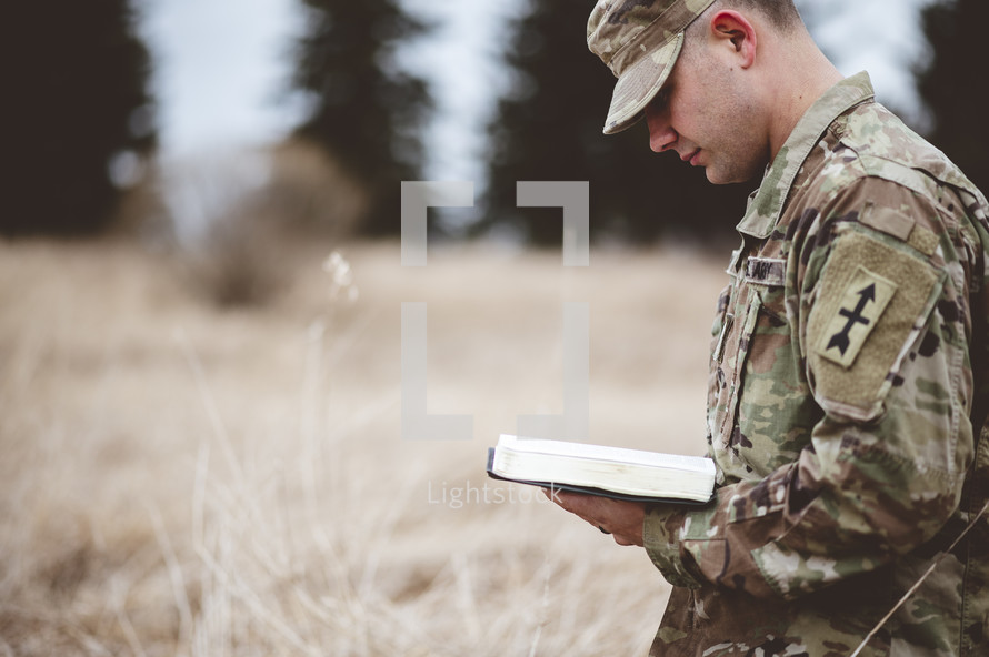 serviceman kneeling in prayer in a field reading a Bible 