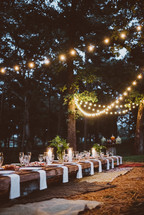 An outdoor table set for a dinner party, lit by strings of lights.