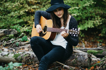 A young woman sits on a tree trunk playing a guitar.