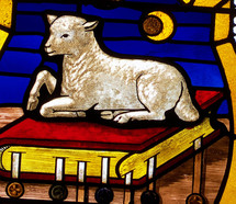 The Lamb of God resting on God's Holy Word stained glass window with red, white, blue, gold, brown colors showing Jesus as the Lamb of God that was slain for the sins of mankind. 