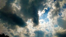 A view of the sun shining through the clouds against a blue sky and gray and silver lined clouds looking like something out of a story book. 