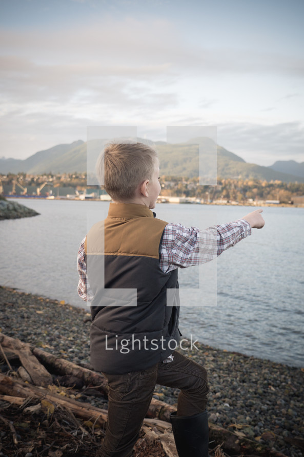 a boy child standing on a shore pointing out over the water 