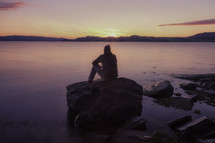 man sitting on a rock watching the sunset over water 