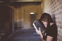 a woman leaning against a brick wall reading a Bible and praying 