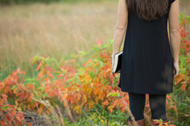 a woman walking in a field holding a Bible 