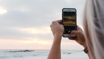 a woman filming the sunset over the ocean with her cellphone 