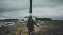 a woman in a hooded sweater walking on the shore near a lighthouse 